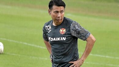 Male soccer player in a dark training shirt standing on a grassy field with hands on hips, rain falling.