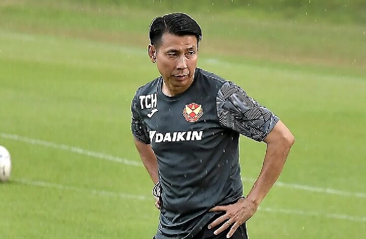 Male soccer player in a dark training shirt standing on a grassy field with hands on hips, rain falling.