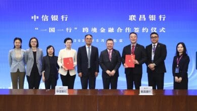 chu kok wei - Wilayah.com.my Eight professionals stand behind a conference table holding red folders at a cross-border banking signing ceremony, in front of a blue banner with Chinese text.