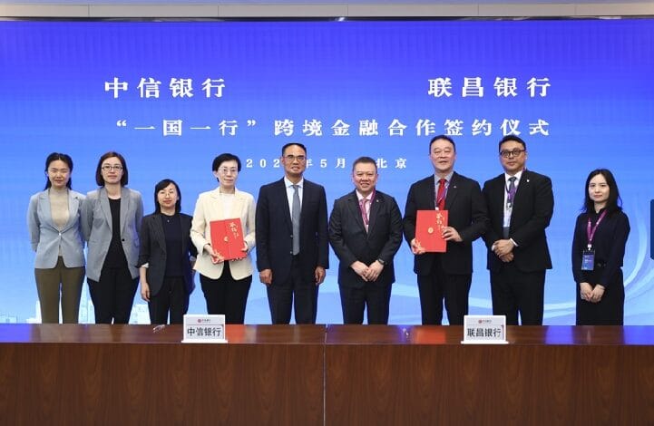 Eight professionals stand behind a conference table holding red folders at a cross-border banking signing ceremony, in front of a blue banner with Chinese text.