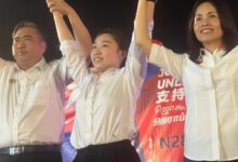 chu poh yee - Wilayah.com.my Three people in white shirts raise their arms together on a stage, smiling.