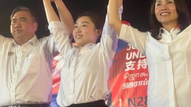 chu poh yee - Wilayah.com.my Three people in white shirts raise their arms together on a stage, smiling.