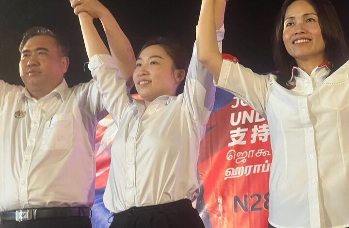 chu poh yee - Wilayah.com.my Three people in white shirts raise their arms together on a stage, smiling.