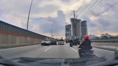 View from a car's windshield on a multi-lane highway with several cars and a motorcycle ahead; construction cranes and tall buildings under a cloudy sky in the distance