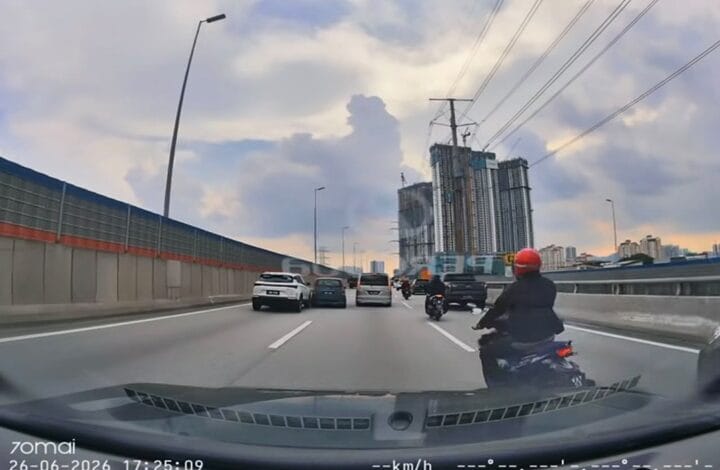 View from a car's windshield on a multi-lane highway with several cars and a motorcycle ahead; construction cranes and tall buildings under a cloudy sky in the distance