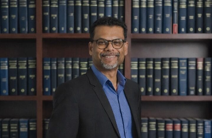 data - Wilayah.com.my Smiling man in a dark blazer and blue shirt stands in a library with wooden bookshelves filled with blue volumes.