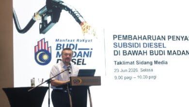 Man at a podium giving a talk, with a large presentation screen and diesel subsidy imagery in the background