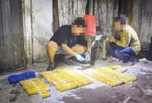 Two people sit in a dim room beside rows of yellow rectangular packages on the floor.