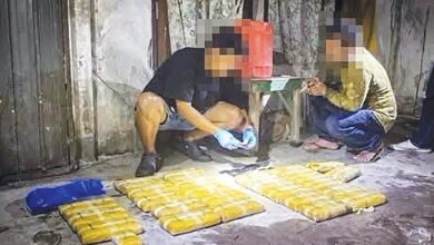 Two people sit in a dim room beside rows of yellow rectangular packages on the floor.