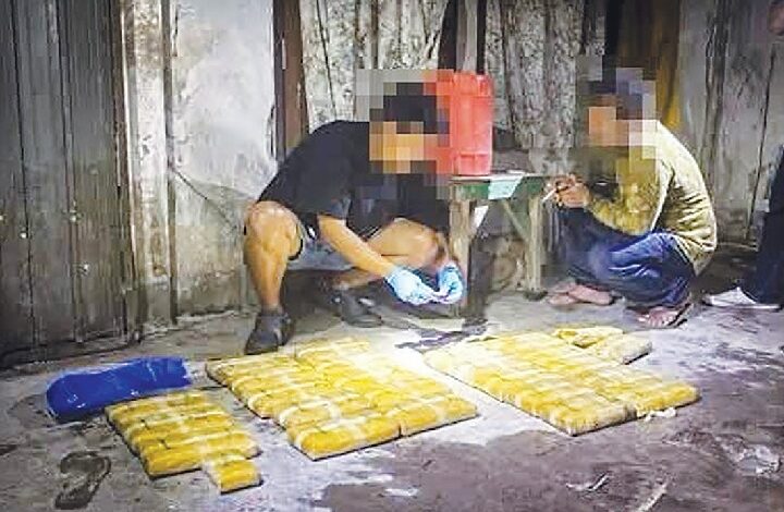 Two people sit in a dim room beside rows of yellow rectangular packages on the floor.