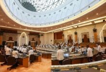 Parliamentary chamber with a large circular skylight, delegates in white shirts seated around curved desks in a grand assembly hall.