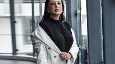 Woman wearing a black blouse and white blazer stands by glass windows in a modern building interior.
