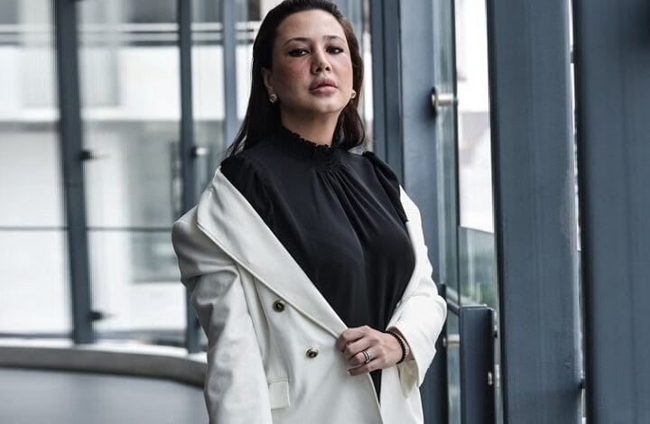 Woman wearing a black blouse and white blazer stands by glass windows in a modern building interior.
