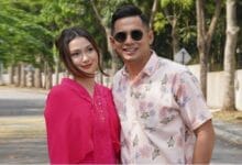 fizo omAR - Wilayah.com.my A couple standing outdoors on a sunny street, smiling at the camera; woman in a bright pink top, man in a light floral shirt with sunglasses.