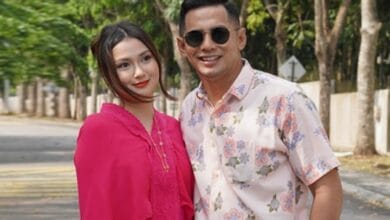 fizo omAR - Wilayah.com.my A couple standing outdoors on a sunny street, smiling at the camera; woman in a bright pink top, man in a light floral shirt with sunglasses.