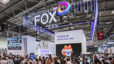 Busy expo floor at the FOX ESS booth with a large illuminated logo, rainbow emblem, and a screen showing a cartoon fox.