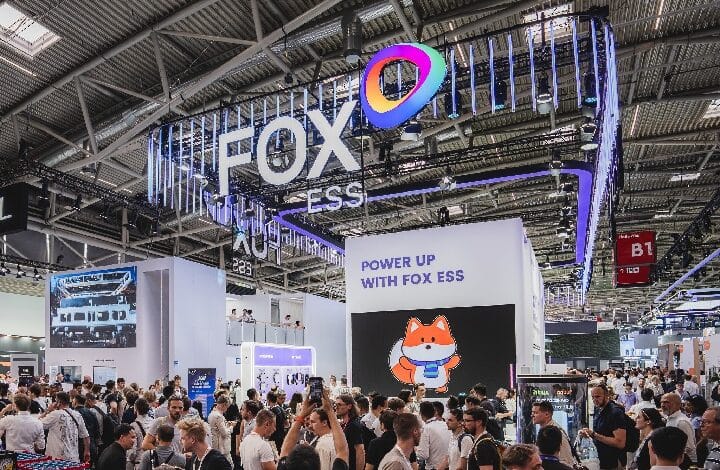 Busy expo floor at the FOX ESS booth with a large illuminated logo, rainbow emblem, and a screen showing a cartoon fox.