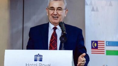 Man in a navy suit and red patterned tie speaks at a podium labeled 'Hotel Royal Signature,' with a microphone and a backdrop featuring flags and banners behind him.