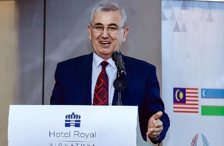 Man in a navy suit and red patterned tie speaks at a podium labeled 'Hotel Royal Signature,' with a microphone and a backdrop featuring flags and banners behind him.