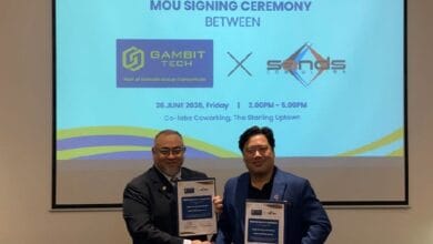 Two men in suits shake hands while holding framed certificates at an MOU signing ceremony; a slide behind displays the event title and partner logos.