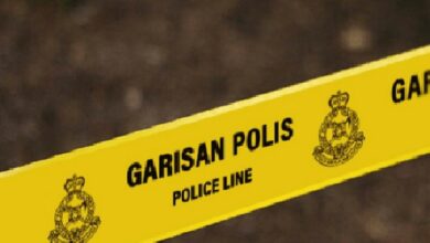 Yellow police line tape marked GARISAN POLIS (POLICE LINE) stretches across the scene with police emblems visible on the sides, a crime-scene barrier.