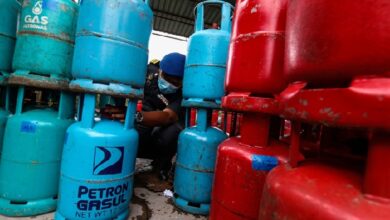 gas - Wilayah.com.my Worker wearing a face mask crouches between blue and red gas cylinders in a storage area, inspecting a valve.