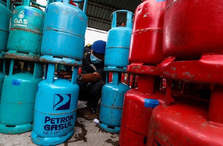 gas - Wilayah.com.my Worker wearing a face mask crouches between blue and red gas cylinders in a storage area, inspecting a valve.