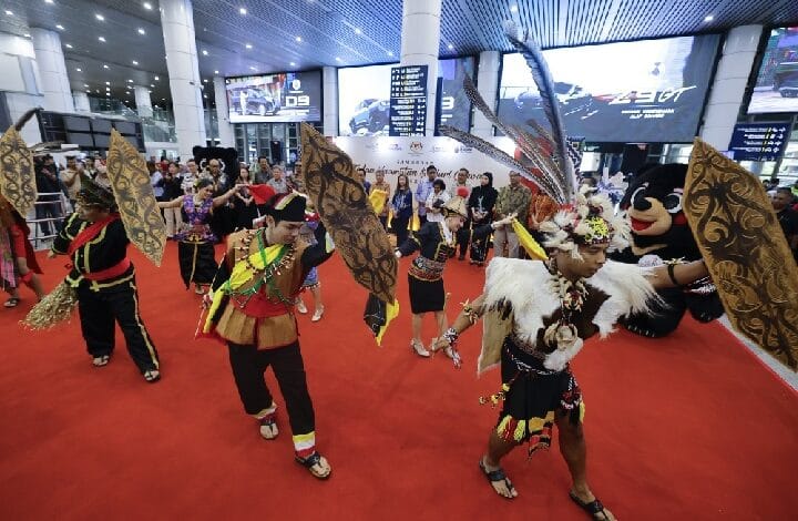 Group of performers in colorful traditional costumes dancing with large decorative shields on a red carpet inside a busy indoor hall.