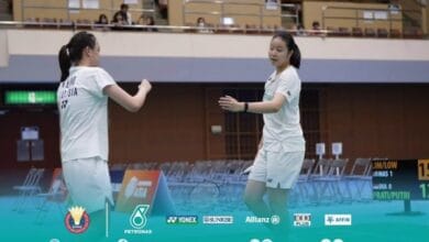 Two female badminton players in white Malaysia jerseys high-fiving on an indoor court with empty stands behind them.