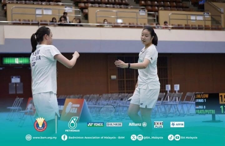 Two female badminton players in white Malaysia jerseys high-fiving on an indoor court with empty stands behind them.
