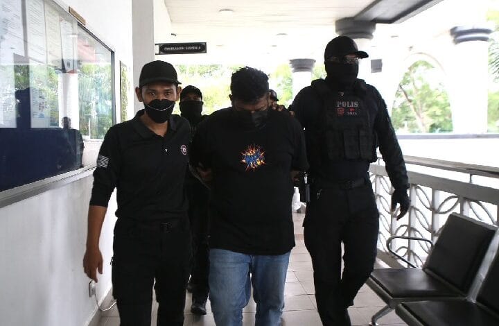 A man in a black T-shirt and jeans is escorted by two masked police officers in black uniforms through a bright corridor, his hands restrained behind his back.
