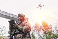 getac zw80 - Wilayah.com.my Soldier in camouflage gear uses a tablet while a quadcopter drone flies overhead in a forested outdoor setting.