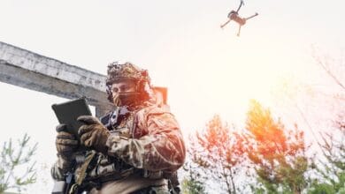Soldier in camouflage gear uses a tablet while a quadcopter drone flies overhead in a forested outdoor setting.