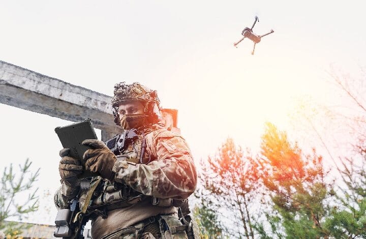 getac zw80 - Wilayah.com.my Soldier in camouflage gear uses a tablet while a quadcopter drone flies overhead in a forested outdoor setting.