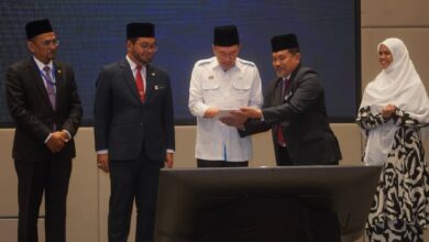 Five officials in suits and a woman in a hijab stand around a large monitor as one man hands a document to another during a formal ceremony.