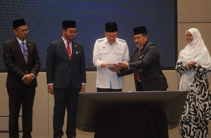 Five officials in suits and a woman in a hijab stand around a large monitor as one man hands a document to another during a formal ceremony.