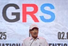 grs - Wilayah.com.my Man in a light pink shirt and cap speaks at a podium with a large GRS logo on a backdrop.