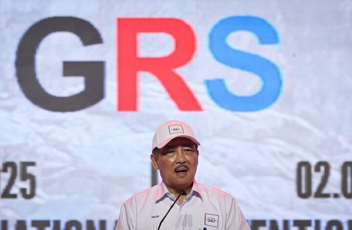 grs - Wilayah.com.my Man in a light pink shirt and cap speaks at a podium with a large GRS logo on a backdrop.
