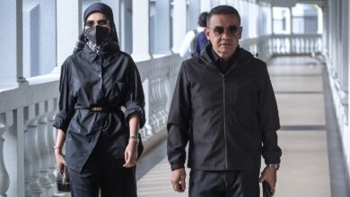 Two adults walk side by side along a sunlit covered walkway: a man in a black jacket and sunglasses, and a woman in a black outfit with a hijab, face mask, and belt, carrying a small purse.