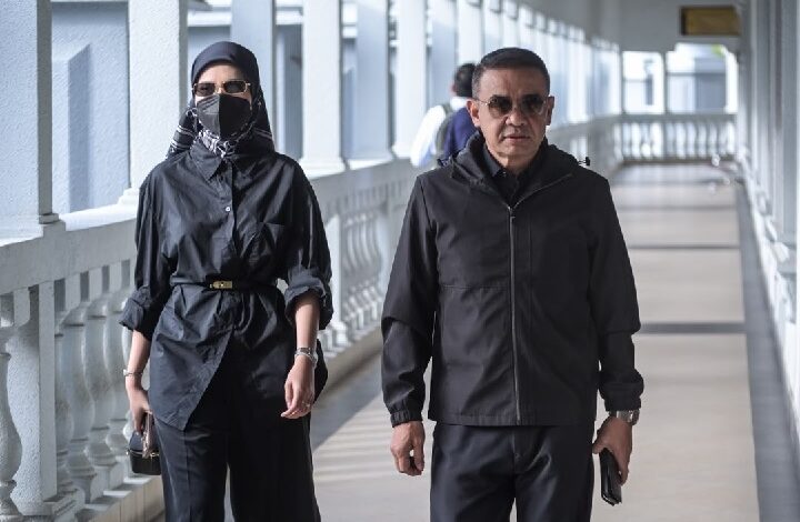 Two adults walk side by side along a sunlit covered walkway: a man in a black jacket and sunglasses, and a woman in a black outfit with a hijab, face mask, and belt, carrying a small purse.