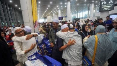 Crowded airport terminal with people hugging and greeting each other, many wearing white caps, surrounded by blue suitcases and rolling bags.