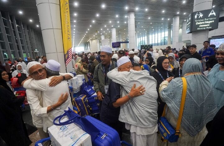 Crowded airport terminal with people hugging and greeting each other, many wearing white caps, surrounded by blue suitcases and rolling bags.