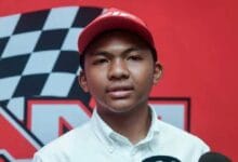Portrait of a young man wearing a red baseball cap and white button-down shirt, posed in front of a red backdrop with a black-and-white checkered race flag design.