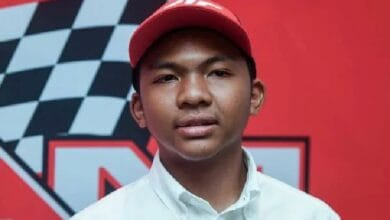 Portrait of a young man wearing a red baseball cap and white button-down shirt, posed in front of a red backdrop with a black-and-white checkered race flag design.