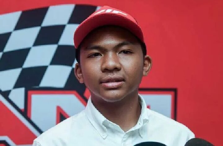 Portrait of a young man wearing a red baseball cap and white button-down shirt, posed in front of a red backdrop with a black-and-white checkered race flag design.