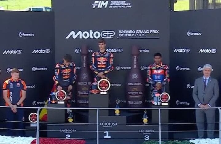 Podium ceremony at a MotoGP event with three riders in racing suits standing on first, second, and third place steps, trophies in hand, and a suited official on the right, against sponsor banners like MotoGP and Brembo in the background.