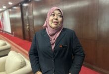 halimaton shaadiah saad - Wilayah.com.my Professional woman in a pink hijab and dark blazer stands in a wood-paneled hallway with a red carpet, wearing a name badge.