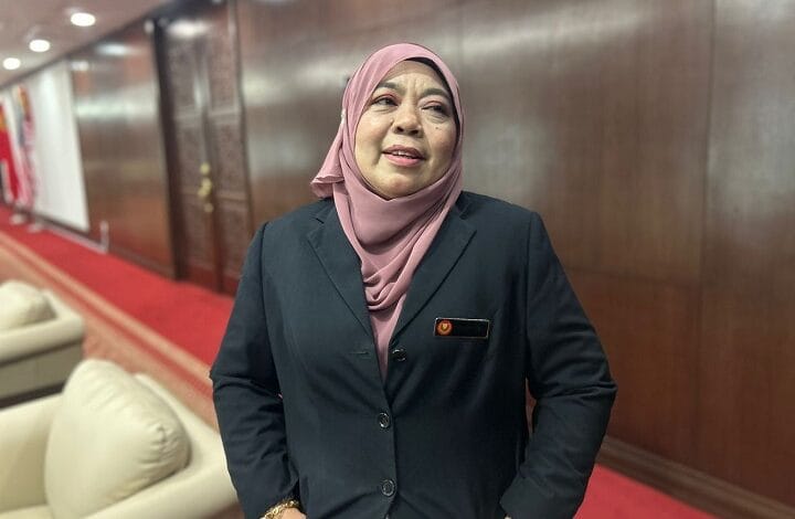 halimaton shaadiah saad - Wilayah.com.my Professional woman in a pink hijab and dark blazer stands in a wood-paneled hallway with a red carpet, wearing a name badge.