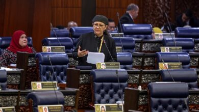 Woman in black attire and hijab speaks at a podium in a formal parliament chamber with blue seats and other officials nearby.