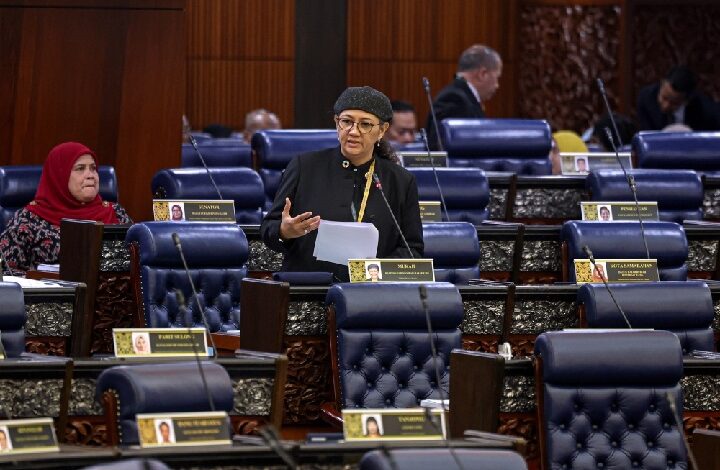 Woman in black attire and hijab speaks at a podium in a formal parliament chamber with blue seats and other officials nearby.